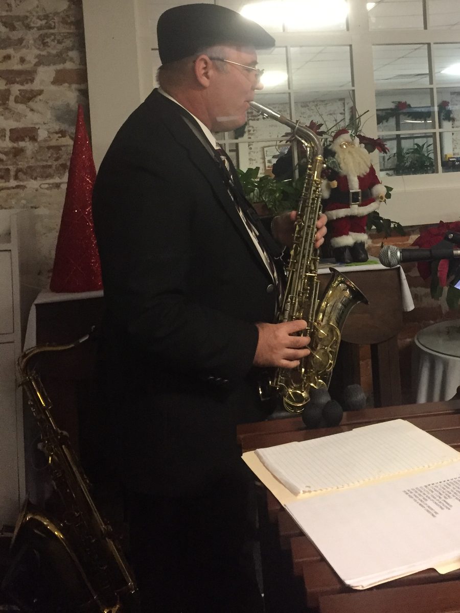 Joey plays alto saxophone in a dark suit and flat cap.